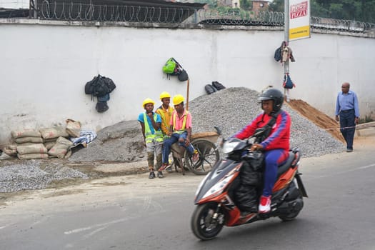 Antananarivo, Madagascar - April 24, 2019: Unknown Malagasy workers in hard hats and reflective jackets standing near gravel heap at construction site next to main road, other man on scooter rides by