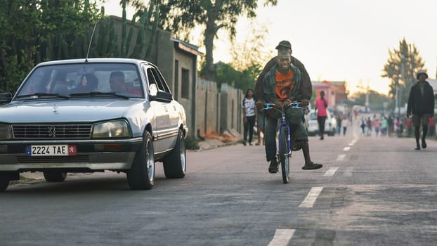Ranohira, Madagascar - April 29, 2019: Two unknown Malagasy locals riding on one bicycle, car next to them, but most people are just walking as they're too poor in this region to afford any transport