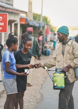 Ranohira, Madagascar - April 29, 2019:  Unknown Malagasy man selling fruit drink to two young girls on street, pouring it directly from sprinkle can to their metal cup. It is cheaper compared to shop