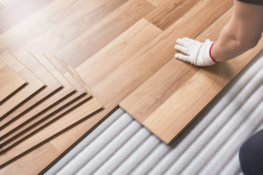 Installing laminated floor, detail on man hand in white glove fitting wooden tile, over white foam base layer