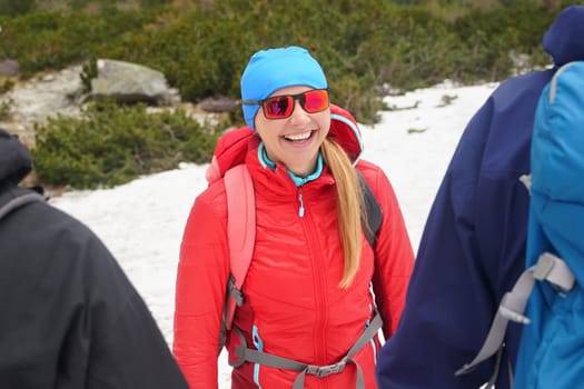 Young woman hiker in red jacket and sunglasses smiling to two other hikers, snow and trees behind her