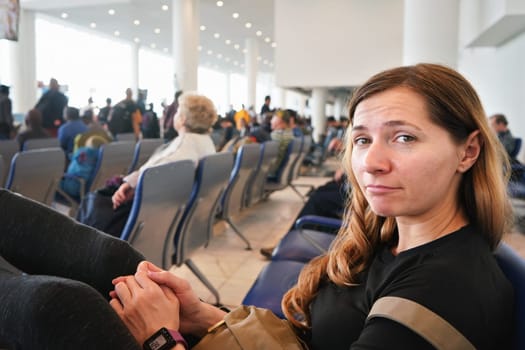 Young woman sitting at airport hall, looking tired after waiting couple of hours for connecting flight in early morning, more blurred passengers in background