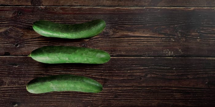 Three green cucumbers laying on dark wooden table, view from above, wide banner, space for text right side