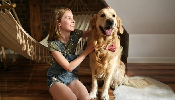 Girl child with golden retriever dog sitting at floor together at home with hammock. Pretty preteen kid hugging purebred doggy pet in loft room
