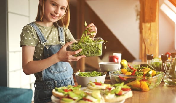 Girl child holding tray with green arugula at kitchen and smiling. Pretty female kid with vegetables and plants preparing vegetarian lunch