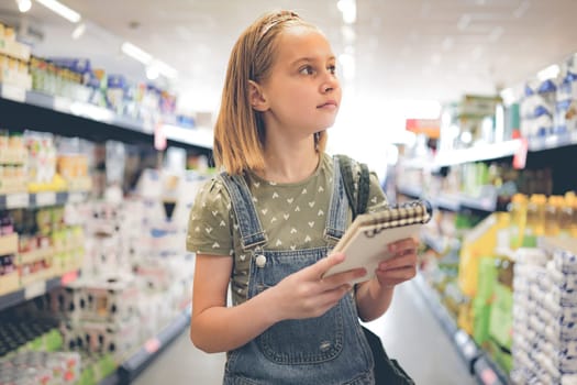 Pretty girl child buying with shopping list in supermarket and looking production shelf. Beautiful female preteen kid in grocery store