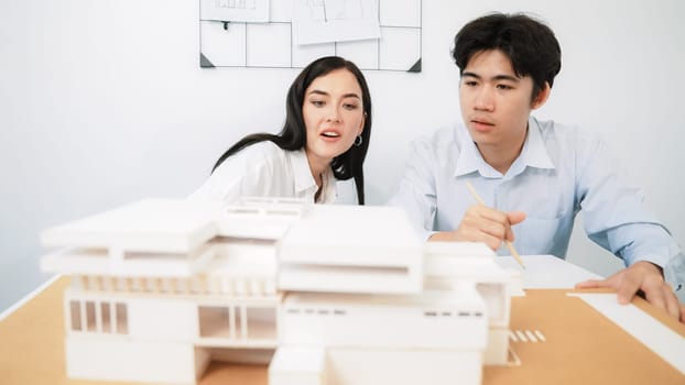 Closeup of skilled architecture engineer team measure and decide house model while working together and discussing about house structure at meeting room with blueprint placed behind. Immaculate.