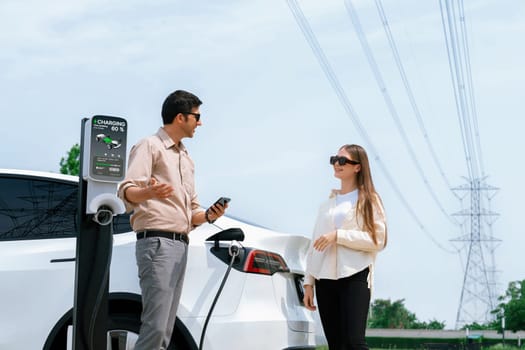 Couple pay for electricity with smartphone while recharge EV car battery at charging station connected to power grid tower electrical as electrical industry for eco friendly car utilization.Expedient