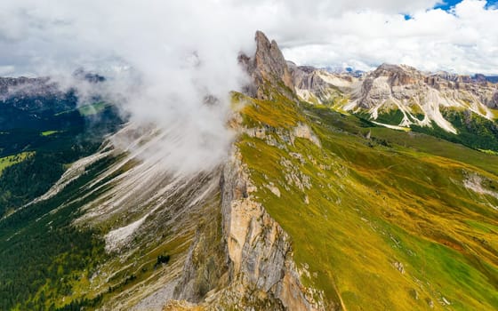 Dense foggy cloud descends on giant Seceda ridgeline with green slopes and bare peak. Cloudy sky above touristic Italian Alps aerial view