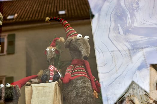 Whimsical New Year's Mouse in Cap and Scarf Stands Behind Store Display. Capture the charm of the holiday season with this delightful image featuring a toy mouse or rat wearing a comical New Year's cap and scarf, playfully standing behind a shop window display. The festive scene adds a touch of whimsy to your creative projects, invoking the spirit of celebration and joy.