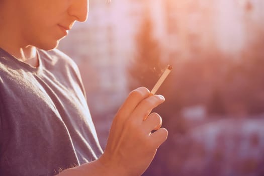 A young man smokes, holds a cigarette in his hand and inhales the smoke while standing at the window early in the morning in the sun, close-up view.