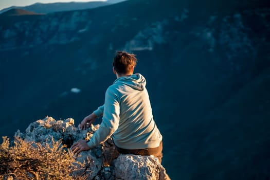 A man sits on a rocky peak and enjoys the mountain landscape at sunset, the traveler discovers trekking, hiking in outdoor adventure, silhouette of a person on the background of nature.