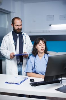 Caucasian doctor and female nurse communicatng in a hospital office. They discuss medical details while using technology such as a tablet and desktop computer for easier research.