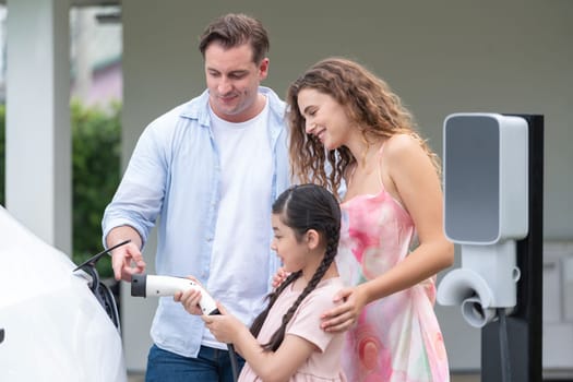 Happy little young girl learn about eco-friendly and energy sustainability as she help her family recharge electric vehicle from home EV charging station. EV car and modern family concept. Synchronos