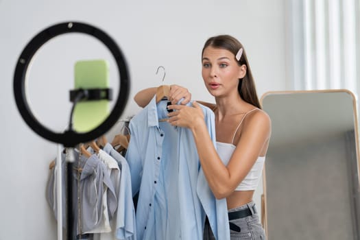 Young woman making fashion video content for social media. Blogger smiles to camera using selfie stick and light ring while making persuasive online clothing sell to audience or followers. Unveiling