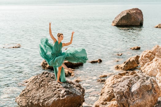 Woman green dress sea. Female dancer in a long mint dress posing on a beach with rocks on sunny day. Girl on the nature on blue sky background