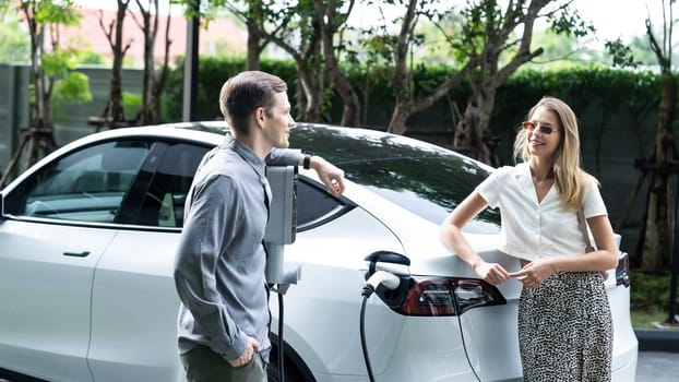 Young couple travel with EV electric car charging in green sustainable city outdoor garden in summer shows urban sustainability lifestyle by green clean rechargeable energy of electric vehicle innards