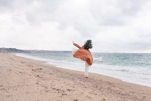 Blond woman Christmas sea. Christmas portrait of a happy woman walking along the beach and holding a Christmas tree on her shoulder. She is wearing a brown coat and a white suit