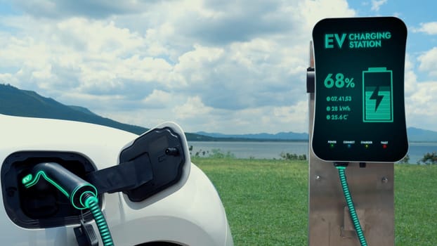 Electric car plugged in with charging station to recharge battery by EV charger cable with nature and lake background. Future innovative ev car and energy sustainability. Peruse