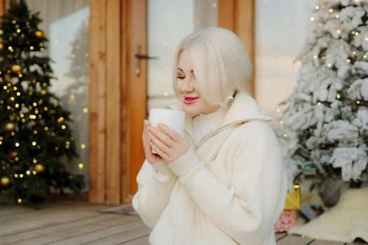 Happy blonde at home at Christmas time, drinking hot beverage from the white mug. Christmas tree in the background. Celebrating Christmas and New Year at home in cozy.