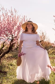 Woman blooming peach orchard. Against the backdrop of a picturesque peach orchard, a woman in a long white dress and hat enjoys a peaceful walk in the park, surrounded by the beauty of nature