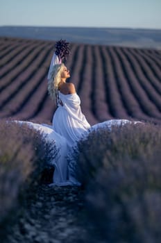 Blonde woman poses in lavender field at sunset. Happy woman in white dress holds lavender bouquet. Aromatherapy concept, lavender oil, photo session in lavender.