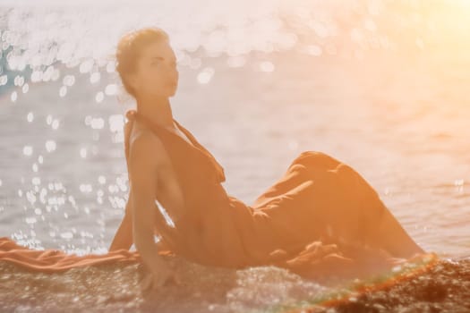 Side view a Young beautiful sensual woman in a red long dress posing on a rock high above the sea during sunrise. Girl on the nature on blue sky background. Fashion photo.