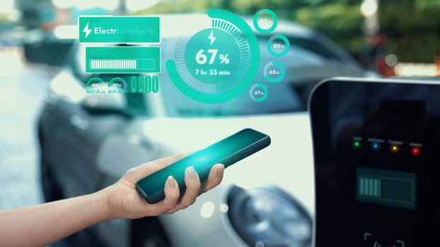Modern businessman check battery status from smartphone mobile 3D hologram while his EV car recharging energy from charging. Innovative technological advancement of future EV automotive tech. Peruse