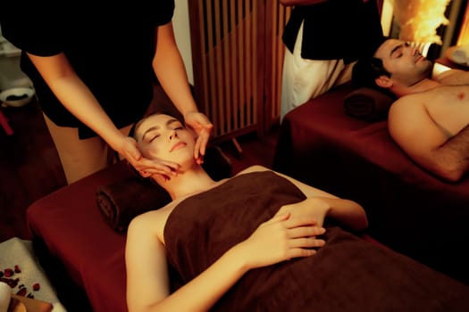 Couple customer enjoying relaxing anti-stress head massage and pampering facial beauty skin recreation leisure in warm candle lighting ambient salon spa in luxury resort or hotel. Quiescent