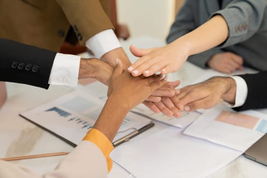 Top view of corporate diverse businesspeople putting hands together on meeting table with document scatter around at business meeting room. Represented unity, cooperation, collaboration. Ornamented.