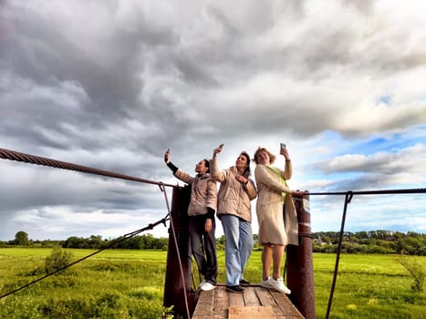 Three funny tourist girls on old bridge take selfies against the background of an alarming dramatic sky with thunderclouds. Middle-aged women having fun outdoors