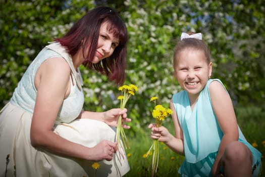 Happy mother and daughter enjoying rest, playing and fun on nature on a green lawn with dandelions and blooming apple tree on background. Woman and girl resting outdoors in summer and spring day