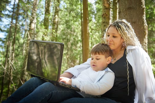 Mother and son with a laptops in the forest in summer. Fat young smart teenage boy and woman working with modern IT technologies in nature