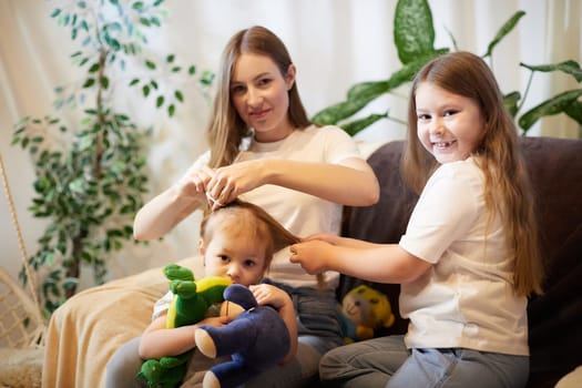 Young loving mom making ponytail to little preschool daughter, adult sister in living room. Mother helping child girl with hairstyle