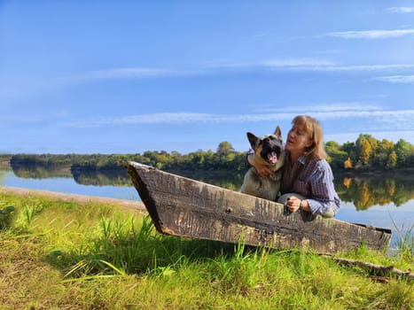 Adult girl with shepherd dog taking selfie on water of river, lake with old boat. Middle aged woman and big pet on nature. Friendship, love, fun, hugs