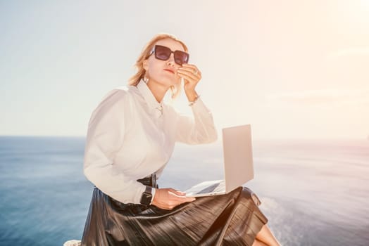 Digital nomad, Business woman working on laptop by the sea. Pretty lady typing on computer by the sea at sunset, makes a business transaction online from a distance. Freelance, remote work on vacation