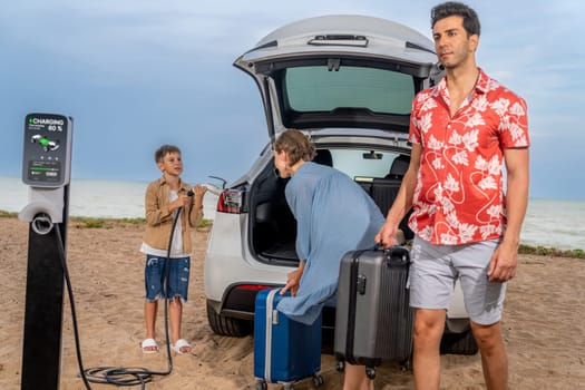 Family vacation trip traveling by the beach with electric car, lovely family taking luggage out while charging EV car battery with clean energy. Alternative family travel by eco-friendly car.Perpetual