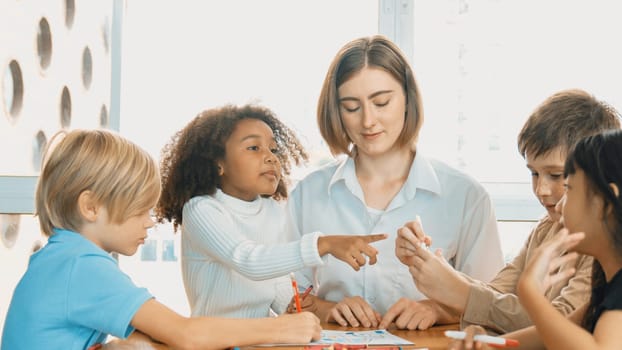 Professional caucasian teacher telling story to diverse student while sitting at table with storybook and colored book. Smart learner listening story while colored picture from instructor. Erudition.