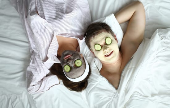 Couple lying on bed with cosmetic mask on face. Husband and wife have fun at home during quarantine. People put cucumbers on their eyes to moisturize their eyelids. Relaxation during self-isolation