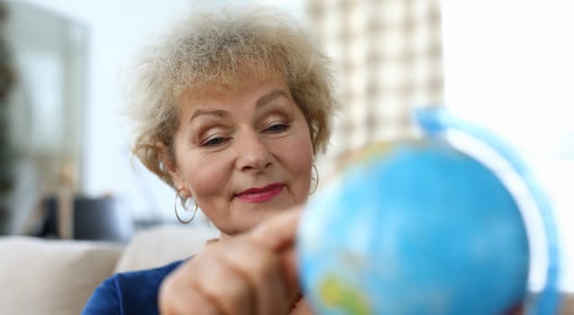 Grandmother looks at globe at home and dreams trip. An elderly woman is considering map states dreaming travels after quarantine. Lonely old woman studying geography during self-isolation apartment