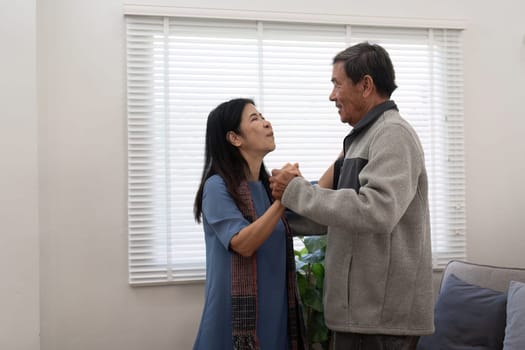 Elderly couple in love looking at each other with tenderness while dancing together in living room at home.