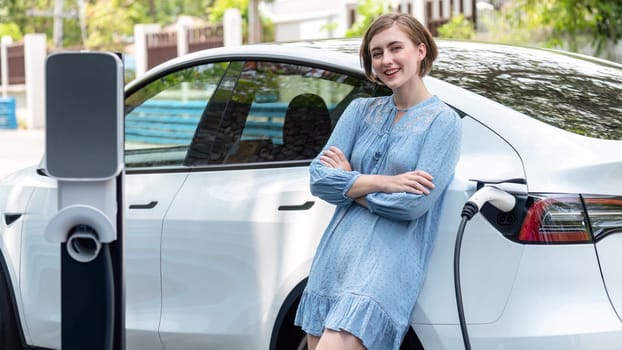 Beautiful young woman recharging her electric car from home EV charging station using alternative energy with net zero emission. Modern young girl charging her vehicle before vacation travel.Perpetual