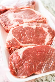 In a sleek, modern white kitchen, a high-quality rib eye steak is carefully being removed from its store packaging. The beautifully marbled steak, soon to be seasoned, promises an upcoming culinary treat as it awaits its turn on the outdoor gas grill.