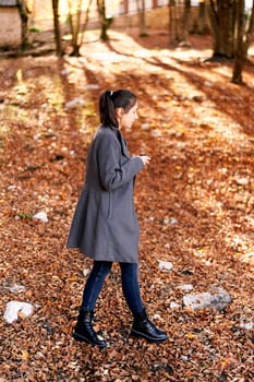 Young woman with a mug walks through the fallen leaves in the park. Side view. High quality photo