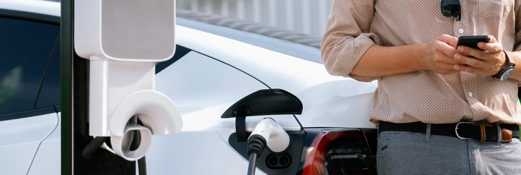 Young man use smartphone to pay for electricity at public EV car charging station at city commercial mall parking lot. Modern environmental and sustainable urban lifestyle with EV vehicle. Expedient