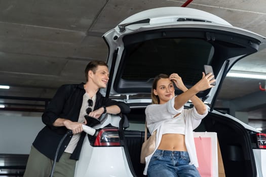 Young couple travel with EV electric car to shopping center parking lot charging in downtown city showing urban sustainability lifestyle by green clean rechargeable energy of electric vehicle innards