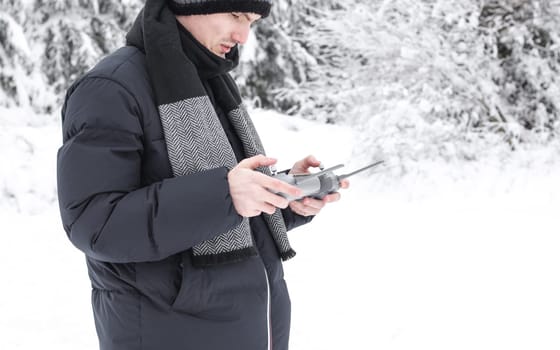 A young handsome caucasian man in a blue down jacket and a gray knitted hat controls a drone through a gray remote control in his hands while standing on a snowy meadow in a winter forest reserve in Belgium, close-up side view. The concept of winter holidays, using technology.