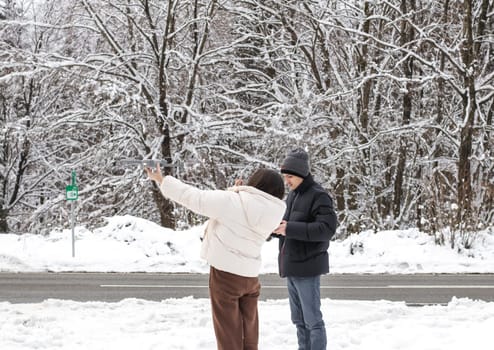 Young beautiful caucasian couple bloggers a guy and a girl launch a gray drone from their hands in a winter forest in belgium to inspect the area, side view close-up.Concept winter vacation.