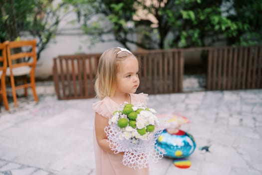 Little girl with a bouquet of flowers stands in the courtyard of the house and looks away. High quality photo