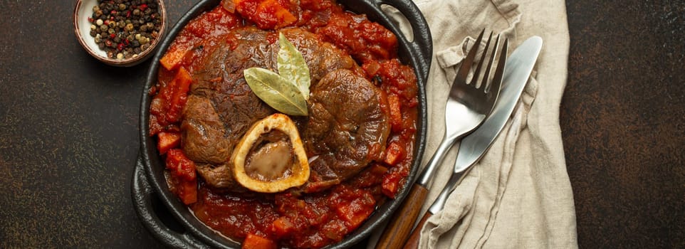 Traditional Italian dish Ossobuco all Milanese made with cut veal shank meat with vegetable tomato sauce served in black casserole pan top view on rustic brown background.